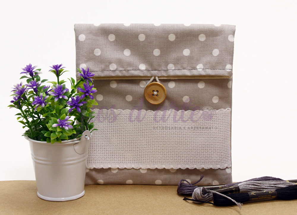 Bolsa de tecido bege com bolinhas brancas e botão de madeira ao lado de vaso branco com flores roxas