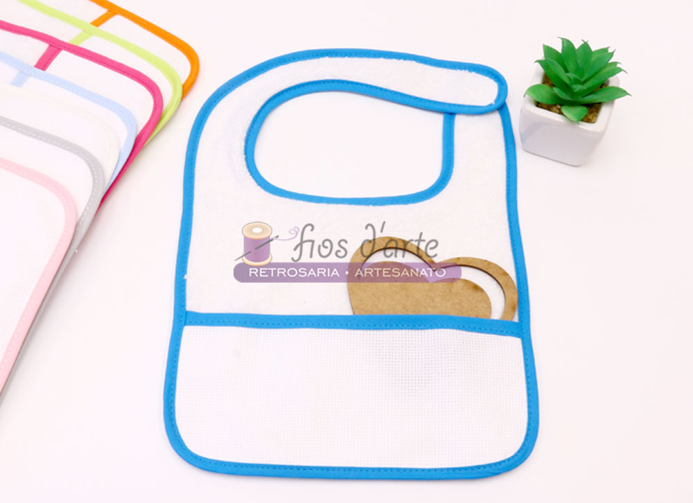 Babete de bebé branco com bordadura azul e bolso frontal, com outros babetes coloridos e planta decorativa ao lado