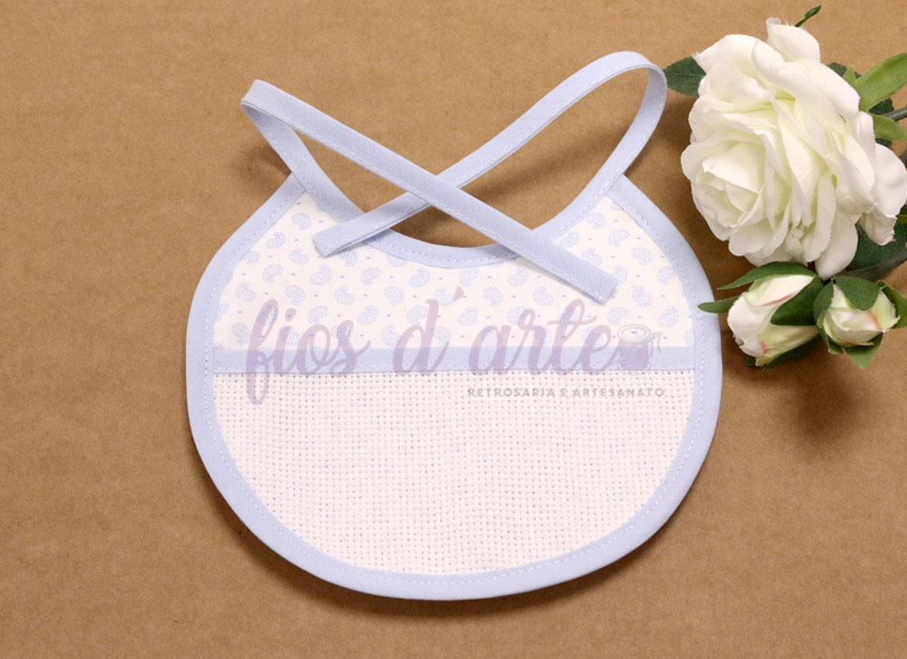 Babete de bebé branco com padrão azul claro e fitas para atar, com flores brancas ao lado