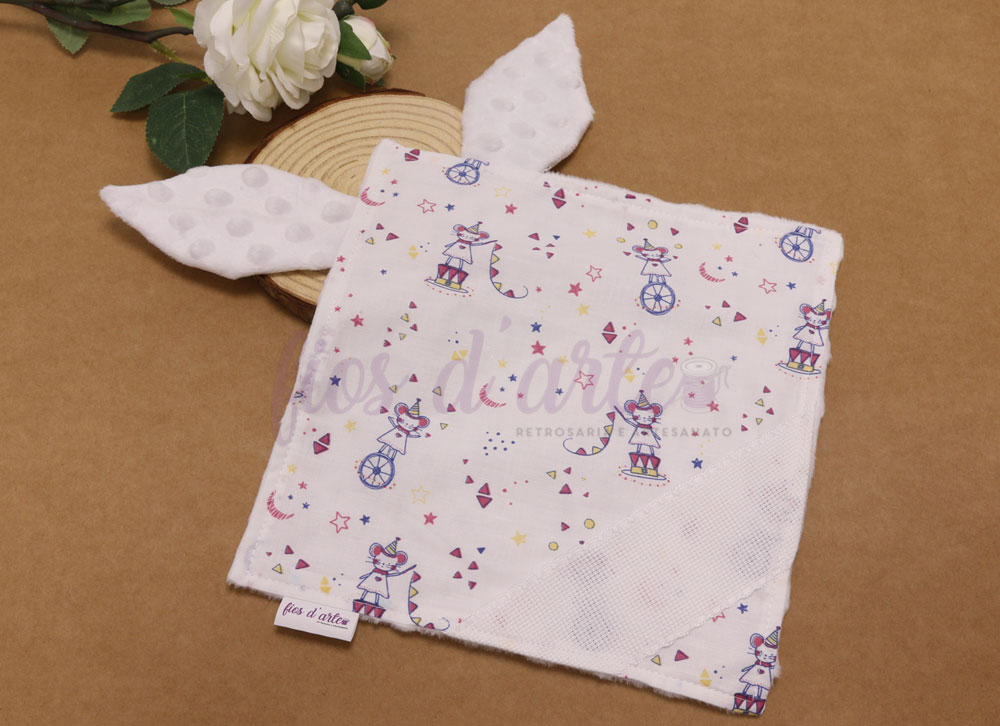 Pano de bebé branco com padrão colorido e orelhas, sobre fundo castanho com flores e madeira.