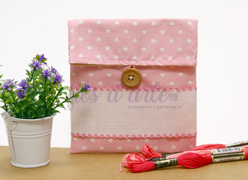 Bolsa rosa com corações brancos, botão de madeira e renda rosa junto a dois novelos de linha vermelha e vaso branco com flores roxas