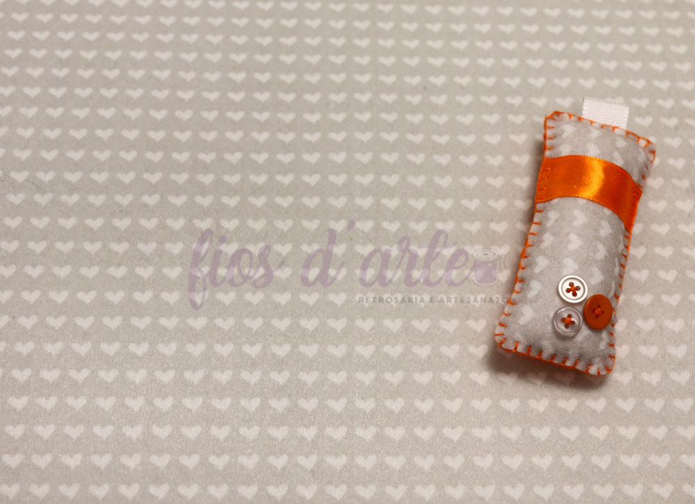 Produto artesanal em feltro cinza com fita laranja e botões sobre tecido bege com corações
