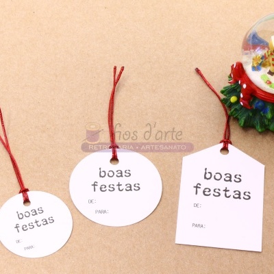 etiquetas de natal brancas com texto 'boas festas' e cordão vermelho ao lado de esfera de neve decorativa