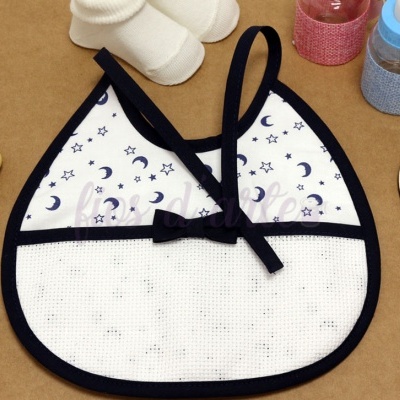 Babete branco com padrão azul de estrelas e luas, laço preto, junto a acessórios de bebé sobre mesa castanha
