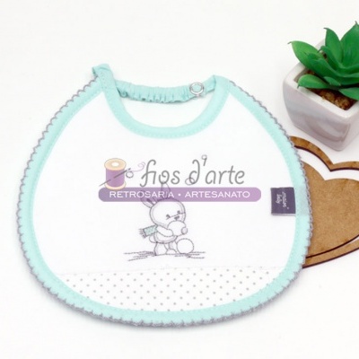 Babete de bebé branco com bordas azuis e estampado de ursinho com texto, junto a vaso com planta verde