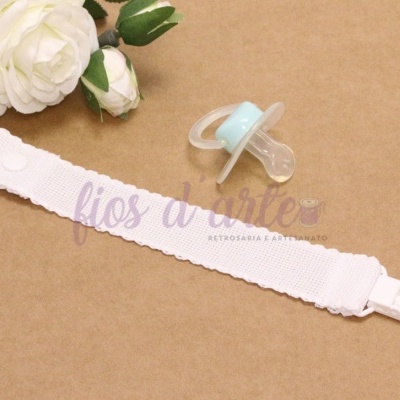 Prendedor de chucha branco com chucha azul e flores brancas no fundo.