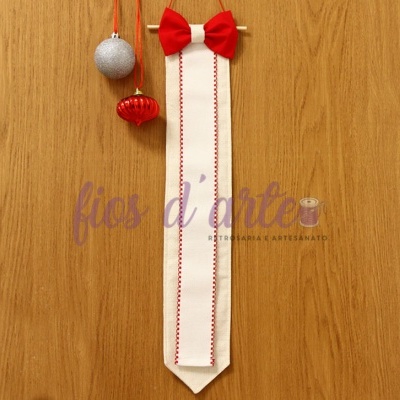 Gravata bege com bordado vermelho e laço vermelho pendurada numa parede de madeira com bolas de Natal ao lado