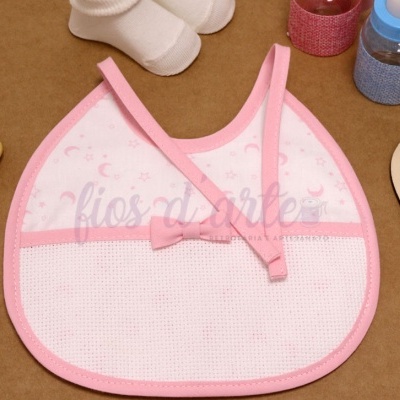 Babete de bebé rosa e branco com padrão de luas e estrelas, em superfície castanha com objetos de bebé