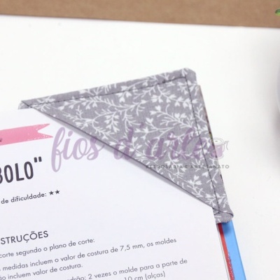 Marcador de livro triangular cinza com padrão branco sobre página de livro e vaso de planta