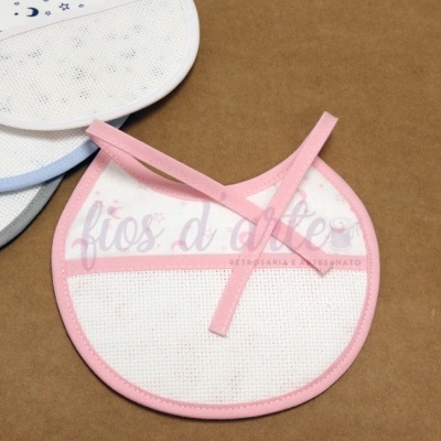 Babeiro branco com bordas rosa e fita para atar, sobre fundo castanho com outros babeiros com bordas em azul, cinza e azul claro