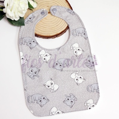 Babete cinzento com padrão de cães, fecho de pressão e etiqueta lateral, sobre base de madeira com flor branca.