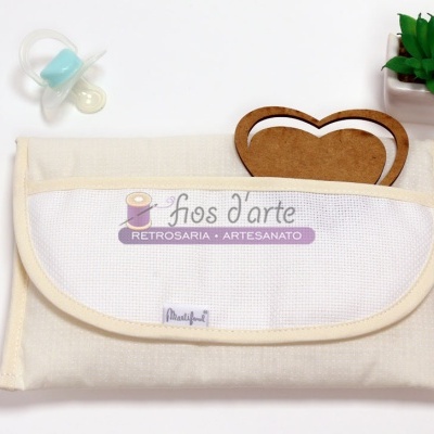 Carteira ou estojo de tecido bege com aba branca e objeto decorativo, chupetas e planta verde ao lado