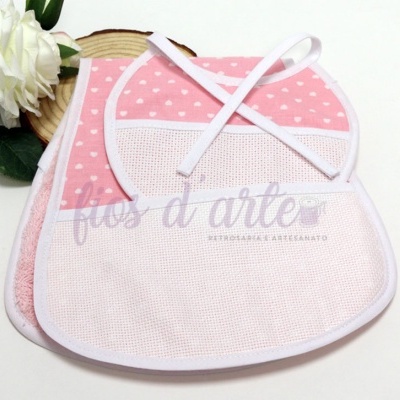 Conjunto de babete rosa com padrão de corações brancos e toalha rosa ao lado de flor branca e madeira