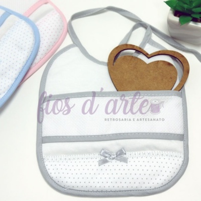 Babete para bebé branco com pontos pretos e laço cinza, dois babetes adicionais com acabamentos azul e rosa, planta verde e moldura de coração de madeira