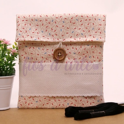 Estojo em tecido bege com padrão floral, botão de madeira e detalhe branco em mesa com planta e linhas