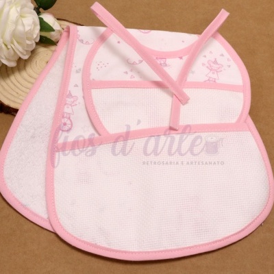 Conjunto de três babetes de bebé brancos com bordas e desenhos cor-de-rosa, sobre fundo castanho com flor branca e base de madeira.