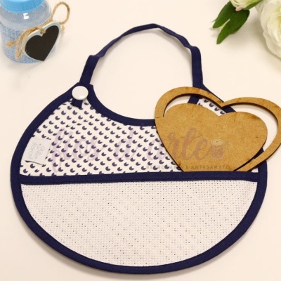 Babete redondo com padrão de luas e pontos, rótulo em forma de coração, flor branca ao lado.