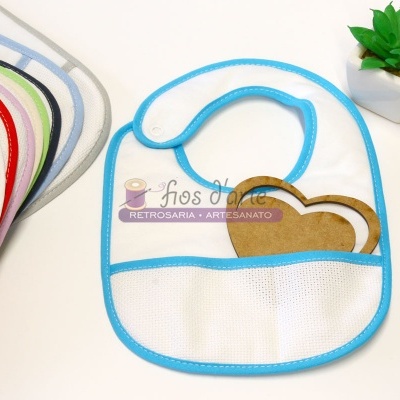 Babete branco com bordas azuis e bolso frontal, várias babetes coloridas ao lado e planta em vaso branco