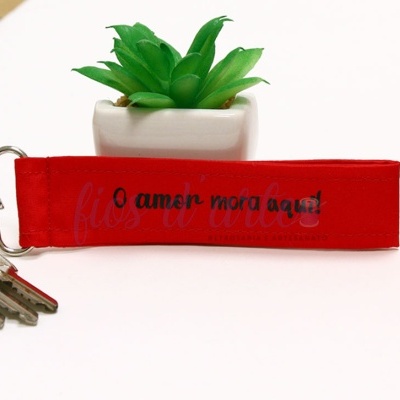 Chaveiro vermelho com texto 'O amor mora aqui' e planta artificial ao fundo
