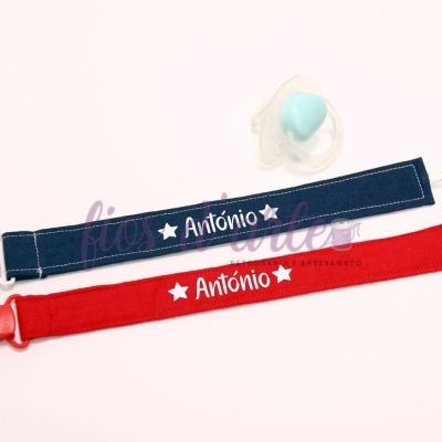 Dois porta-chupetas de tecido azul escuro e vermelho com nome 'António' e estrelas brancas, pacificador azul e transparente