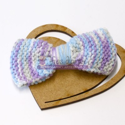 Laço de cabelo tricotado azul, branco e lilás sobre coração de madeira.