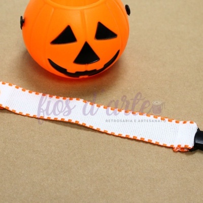 Pulseira para bebé branca com bordas laranja e fecho preto em superfície castanha ao lado de balde de abóbora laranja de Halloween