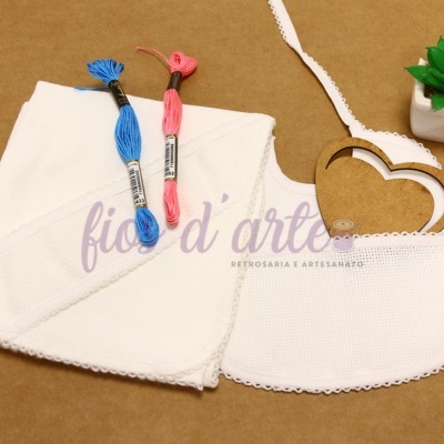 Conjunto de toalha e babete brancos com bordado e novelos de linha azul e rosa, suporte de madeira em forma de coração e planta pequena em vaso branco.