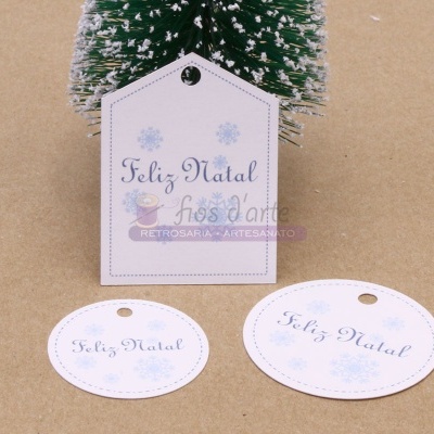 Etiqueta branca em formato de casa com texto 'Feliz Natal' pendurada numa árvore de Natal miniatura com neve artificial