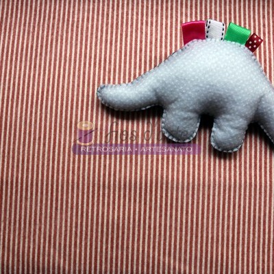 Pelúcia artesanal branca em forma de dinossauro com fitas coloridas sobre tecido listrado vermelho e branco
