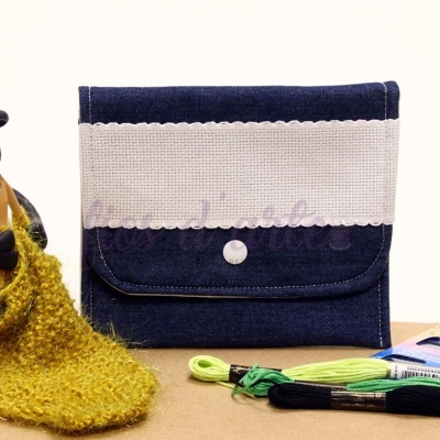 Carteira azul escura com aba cinzenta, fecho de botão, rodeada por materiais de tricô e figura de ovelha