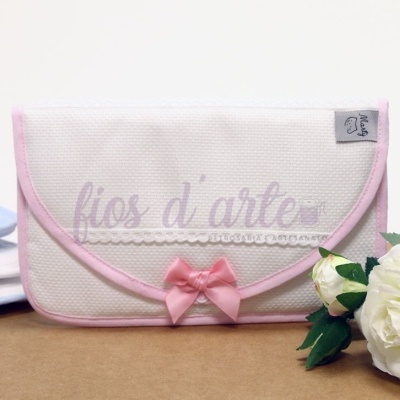 Estojo branco com laço cor-de-rosa, decorado com renda, ao lado de rosas brancas e livros azuis.