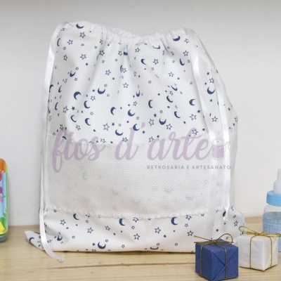Saco de tecido branco com estampado azul de luas e estrelas, com cordões de cetim, sobre mesa de madeira com acessórios infantis