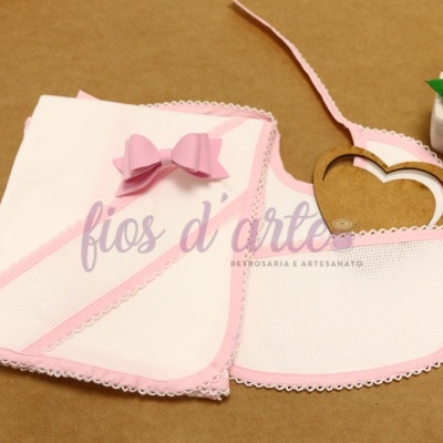 Manta branca com borda e laço rosa e babete branco com borda rosa ao lado de planta verde