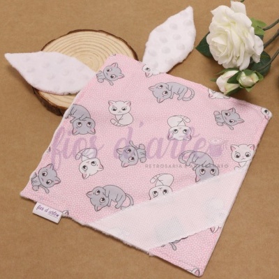 Manta bebé rosa com padrão de gatos e orelhas brancas felpudas junto a flores brancas e base de madeira