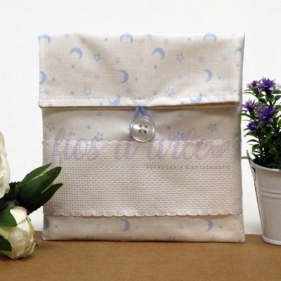 Bolsa de tecido com padrão de luas e estrelas azuis em fundo bege, detalhe em renda branca, entre flores artificiais branca e púrpura.