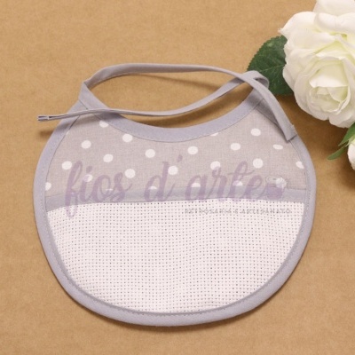 Babete cinzento claro e branco com bolinhas e tecido de tela, com fitas para atar, sobre fundo castanho com flor branca ao lado.