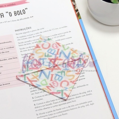 Livro aberto com instruções em português e suporte de tecido colorido com letras em cima da página, vaso branco com planta ao lado