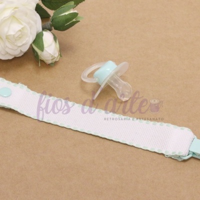 Chupeta transparente e azul clara com suporte de tecido branco e azul colocado sobre fundo bege com flores brancas