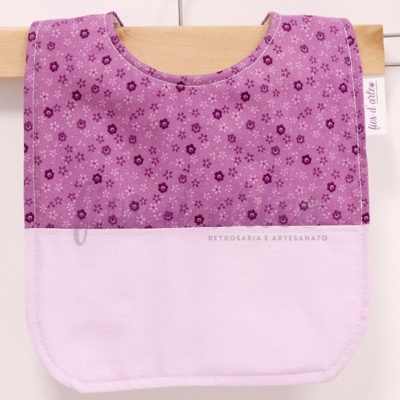 Babete lilás com padrão de flores e parte inferior branca, pendurado numa roupa de madeira.