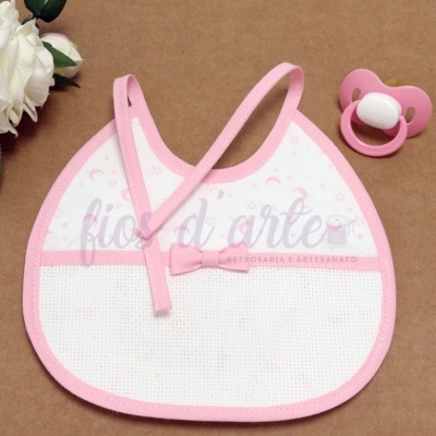 Babete de bebé branco e cor-de-rosa com laço e chupeta ao lado sobre fundo castanho