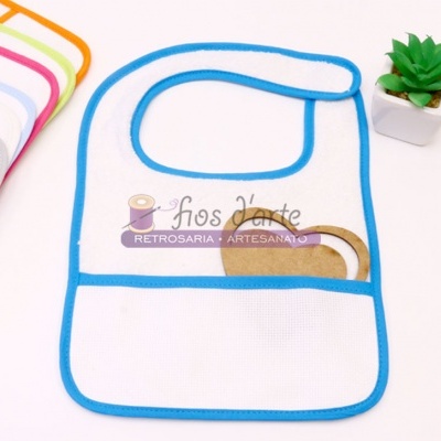 Babete de bebé branco com bordadura azul e bolso frontal, com outros babetes coloridos e planta decorativa ao lado