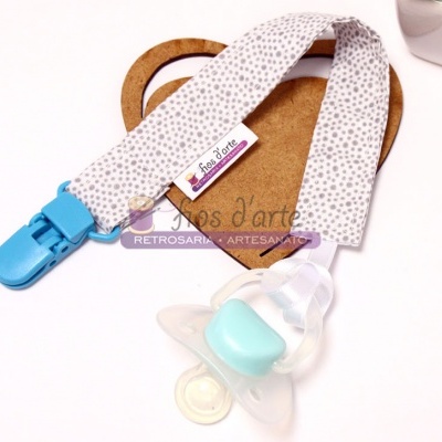 Chupeta com cinto de tecido branco com pontos cinzentos e clip azul sobre suporte de cartão castanho em forma de coração