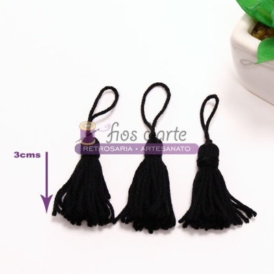 Três borlas pretas pequenas com alça em fundo branco, ao lado de vaso com planta verde