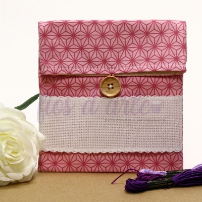 Estojo de tecido rosa com botão de madeira e faixa bordada, com flor branca e novelos de linha roxa ao lado.