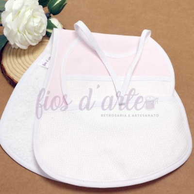 Dois babetes de bebé em algodão, um rosa claro e branco, outro branco, numa superfície castanha com flor e base de madeira.