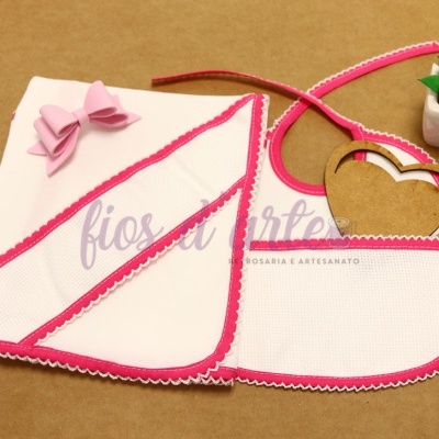 Toalha de banho e babete infantil brancos com acabamento rosa e laço, junto a pequeno vaso de planta verde.