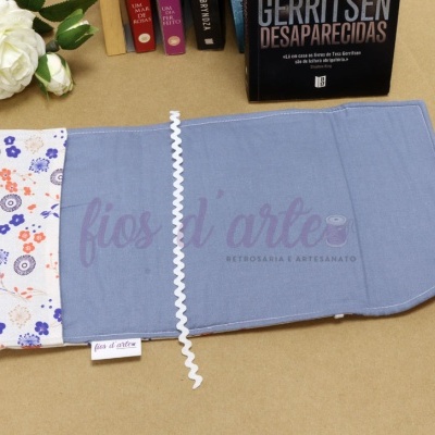 Estojo de tecido azul e branco com padrão floral e fita branca sobre mesa bege, junto a livro e flor branca