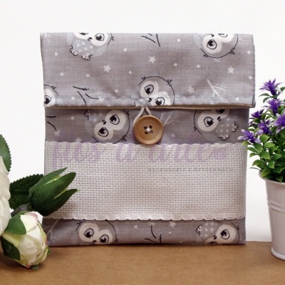 Estojo cinza com padrão de corujas e botão de madeira ao centro, rodeado por flores brancas e lilases.