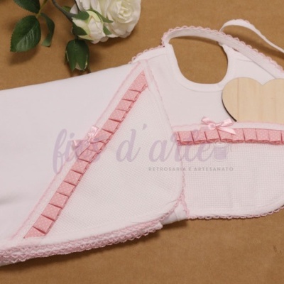 Manta e babete de bebé brancos com detalhes rosa decorativos sobre fundo castanho claro com flores brancas.