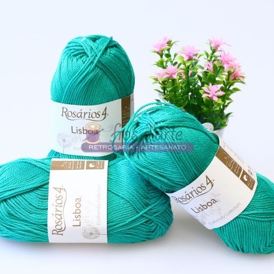 Quatro novelos de lã verde-turquesa Rosários 4 Lisboa com rótulos brancos ao lado de vaso com flores rosas